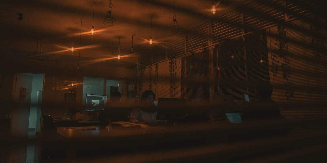 A dim open-plan office at night seen through half-closed blinds, warm pendant lights over occupied desks.