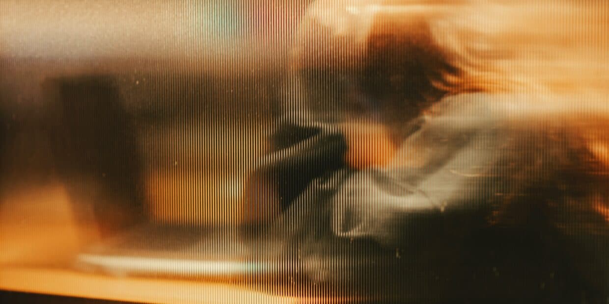 A figure at a desk seen through ribbed glass, smeared by motion into an unreadable shape.